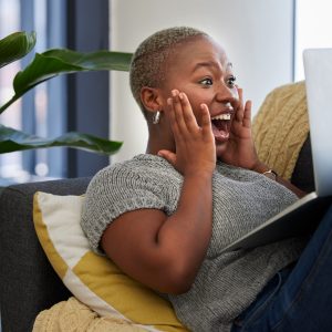 Laptop, surprise and excited black woman on sofa shocked with win of digital lottery, competition o